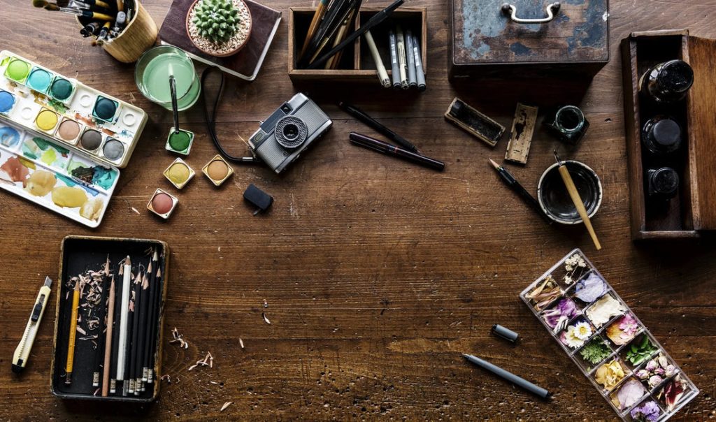 A cluttered artistic workspace with paints, brushes, and tools on a wooden table
