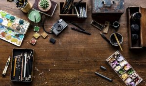 A cluttered artistic workspace with paints, brushes, and tools on a wooden table